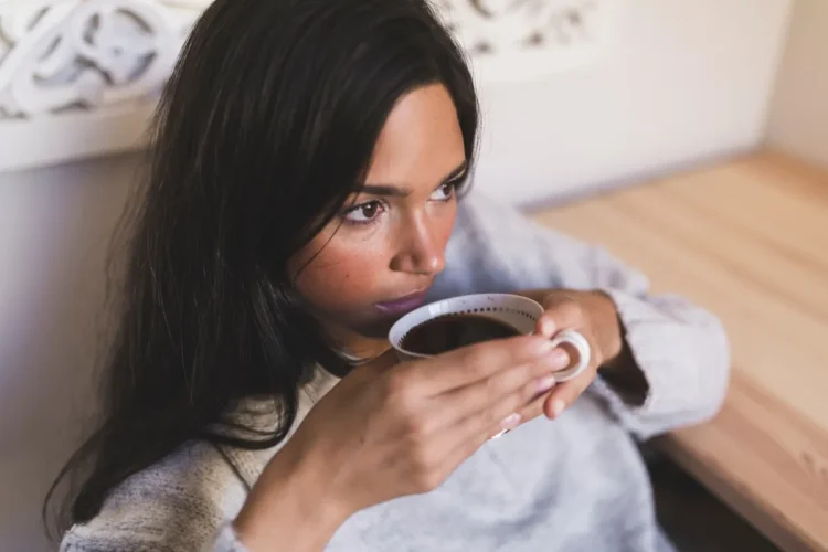 O que acontece com o seu corpo se você beber café de estômago vazio todos os dias? 1 Mulher bebendo café com expressão pensativa, refletindo sobre os efeitos de beber café de estômago vazio.
