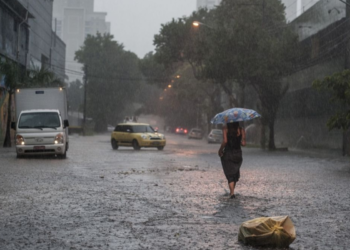 Mulher caminha com guarda-chuva em rua alagada sob chuva intensa, carros ao fundo e sacola no chão.