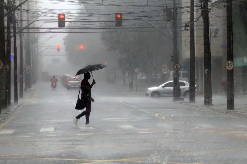 Alerta: Previsão do tempo indica temporais e chuvas fortes para várias regiões brasileiras 2 Homem atravessando rua sob chuva forte e neblina em centro urbano, carregando guarda-chuva preto. Semáforos vermelhos e carros ao fundo. Alerta de temporais no Brasil.