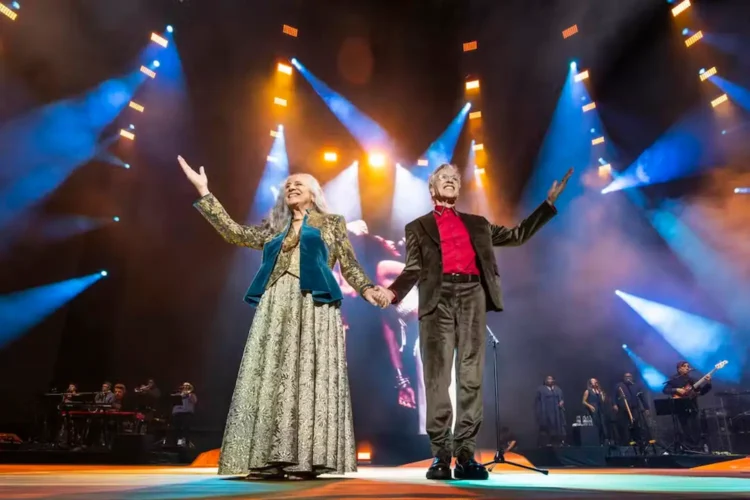 Caetano Veloso e Maria Bethânia conquistam o Grammy 2026 na categoria música global 1 Dois artistas brasileiros se apresentam juntos em um palco iluminado, de mãos dadas, durante um espetáculo musical com banda ao fundo.
