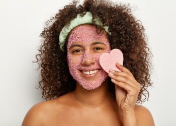 Mulher com esfoliante rosa no rosto segura esponja de coração e sorri para a câmera