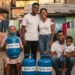 Família reunida na rua sorrindo com botijões de gás azul do programa Vale Recarga Gás do Povo