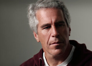 Financista Jeffrey Epstein, um homem de pele clara e cabelos grisalhos curtos, vestindo um moletom bordô sobre uma camiseta branca. Ele tem uma expressão séria e olha levemente para o lado. O fundo é cinza neutro e desfocado.