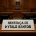 Imagem de uma placa branca sobre uma mesa de madeira em um tribunal, com os dizeres 'Sentença de Hytalo Santos' em letras pretas. Ao fundo, é possível ver cadeiras e o ambiente característico de uma sala de audiência judicial.