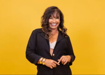 A atriz Solange Couto sorri frontalmente contra um fundo amarelo vibrante. Ela possui cabelos castanhos ondulados com luzes, veste um blazer preto aberto sobre um top de pérolas brancas e usa diversos braceletes dourados no pulso direito.