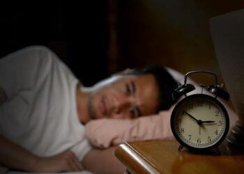 Homem acordado à noite, deitado na cama, olhando para o relógio despertador em um ambiente escuro.