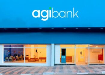 Fachada externa de uma agência do Agibank durante o dia. A imagem mostra o letreiro com o logotipo "agibank" em branco sobre um painel azul vibrante, acima de amplas vitrines de vidro que revelam o interior da unidade.