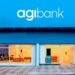 Fachada externa de uma agência do Agibank durante o dia. A imagem mostra o letreiro com o logotipo "agibank" em branco sobre um painel azul vibrante, acima de amplas vitrines de vidro que revelam o interior da unidade.