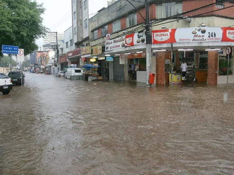 Rua comercial alagada em Juiz de Fora com água barrenta atingindo a metade da altura das portas de uma loja vermelha e encobrindo pneus de carros estacionados ao fundo.