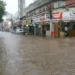 Rua comercial alagada em Juiz de Fora com água barrenta atingindo a metade da altura das portas de uma loja vermelha e encobrindo pneus de carros estacionados ao fundo.
