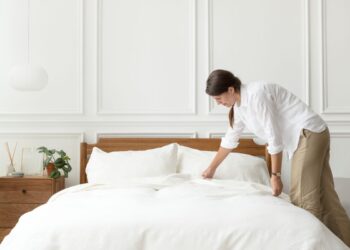 Uma mulher de camisa branca e calça bege inclina-se para arrumar uma cama de casal com lençóis brancos impecáveis. O quarto possui paredes brancas com molduras clássicas, uma mesa de cabeceira de madeira com uma planta pequena e uma luminária pendente moderna à esquerda.