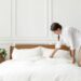 Uma mulher de camisa branca e calça bege inclina-se para arrumar uma cama de casal com lençóis brancos impecáveis. O quarto possui paredes brancas com molduras clássicas, uma mesa de cabeceira de madeira com uma planta pequena e uma luminária pendente moderna à esquerda.