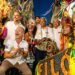 Grupo de pessoas fantasiadas sorrindo e celebrando em rua decorada durante o Pré-Carnaval.