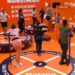 Competidores em pé sobre plataformas circulares laranja, segurando barras, olhos atentos ao centro da arena.