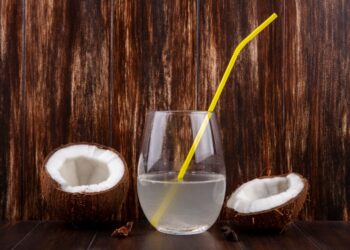 Um copo de vidro transparente com água de coco e um canudo amarelo, posicionado entre duas metades de um coco seco sobre uma mesa de madeira escura com fundo rústico.