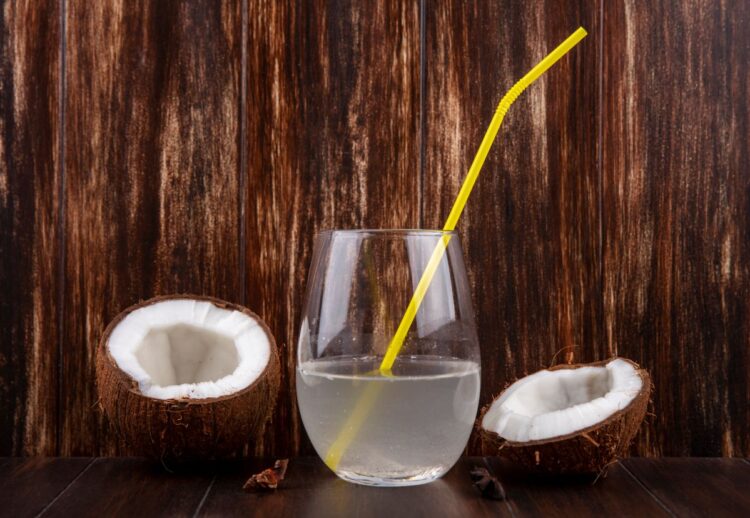 Um copo de vidro transparente com água de coco e um canudo amarelo, posicionado entre duas metades de um coco seco sobre uma mesa de madeira escura com fundo rústico.
