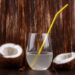 O que acontece se você beber água de coco em jejum? O efeito real no corpo após um dia de exageros 15 Um copo de vidro transparente com água de coco e um canudo amarelo, posicionado entre duas metades de um coco seco sobre uma mesa de madeira escura com fundo rústico.