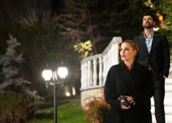 Uma mulher loira com expressão de surpresa veste um sobretudo preto elegante à noite. Atrás dela, um homem de terno risca de giz desce uma escadaria branca iluminada em um jardim luxuoso.