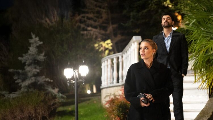 Netflix: 5 séries turcas muito boas que valem a pena assistir 1 Uma mulher loira com expressão de surpresa veste um sobretudo preto elegante à noite. Atrás dela, um homem de terno risca de giz desce uma escadaria branca iluminada em um jardim luxuoso.