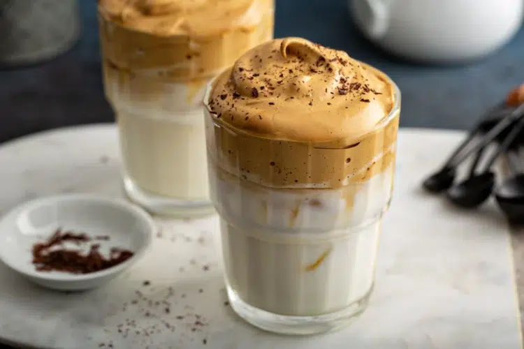 O café cremoso de 3 ingredientes que fica igual ao de cafeteria chique 2 Dois copos de café cremoso com espuma dourada, raspas de chocolate no topo e leite na base, com prato de raspas de chocolate ao lado.