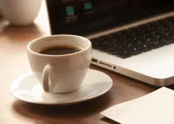 Xícara de café preto sobre pires branco em mesa de madeira ao lado de notebook, representando a rotina de café no ambiente de trabalho.