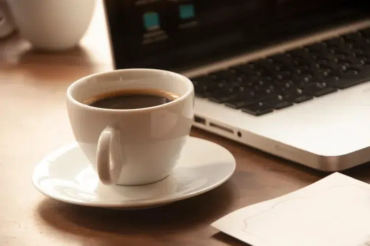 Pessoas que tomam café no trabalho: O que a ciência diz sobre a produtividade delas 1 Xícara de café preto sobre pires branco em mesa de madeira ao lado de notebook, representando a rotina de café no ambiente de trabalho.