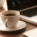 Xícara de café preto sobre pires branco em mesa de madeira ao lado de notebook, representando a rotina de café no ambiente de trabalho.