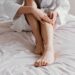 Close-up das pernas de uma pessoa sentada em uma cama com lençóis claros, usando um roupão branco. Suas mãos estão pressionando a panturrilha esquerda, simulando um movimento de massagem para aliviar a dor de uma câimbra noturna. Ao lado, sobre a cama, aparece parte de um frasco de creme, sugerindo o cuidado com a musculatura.