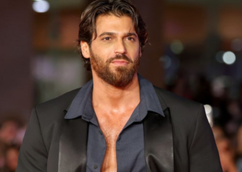 Homem de barba e cabelo castanho veste terno preto e camisa azul em ambiente sofisticado