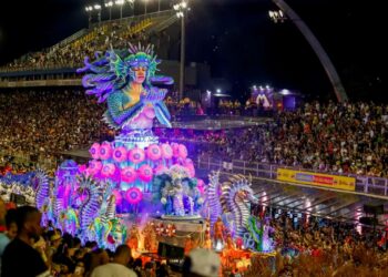 Imagem noturna do Sambódromo do Anhembi em São Paulo durante o Carnaval. Um suntuoso carro alegórico em tons de azul e roxo apresenta uma escultura gigante de uma sereia com as mãos postas, cercada por cavalos-marinhos e iluminação neon vibrante.