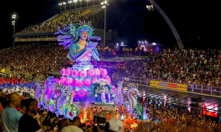 Carnaval SP 2026: confira a programação completa dos desfiles das escolas de samba 1 Imagem noturna do Sambódromo do Anhembi em São Paulo durante o Carnaval. Um suntuoso carro alegórico em tons de azul e roxo apresenta uma escultura gigante de uma sereia com as mãos postas, cercada por cavalos-marinhos e iluminação neon vibrante.