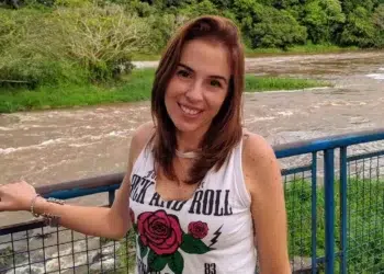 Mulher de cabelos castanhos sorrindo, em ponte azul sobre rio, com camiseta branca, ao ar livre.