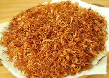 Pilha de anéis de cebola bem finos e fritos até atingirem um tom dourado intenso, repousando sobre uma folha de papel toalha branco. As rodelas exibem uma textura leve e sequinha, típica do empanamento com amido de milho.
