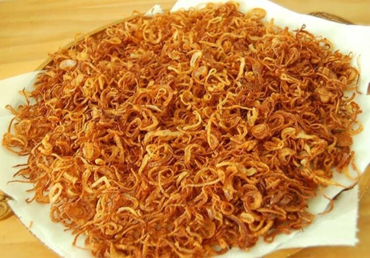 Revelado: O segredo da cebola crocante de hamburgueria 1 Pilha de anéis de cebola bem finos e fritos até atingirem um tom dourado intenso, repousando sobre uma folha de papel toalha branco. As rodelas exibem uma textura leve e sequinha, típica do empanamento com amido de milho.