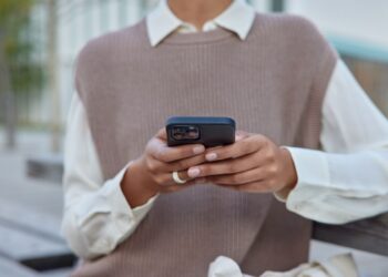 Close-up das mãos de uma pessoa segurando um celular com capa preta, enquanto utiliza os polegares para navegar no aparelho. O fundo está desfocado em tons de marrom e verde, com foco total na interação do usuário com o dispositivo móvel para limpar a memória.