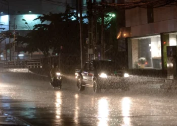 Carros e motocicleta trafegam sob chuva forte à noite em avenida urbana durante temporal.