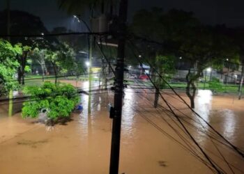 Vista aérea noturna de uma longa avenida urbana totalmente submersa por água barrenta, com postes de luz acesos refletindo na inundação e silhuetas de casas ao fundo.