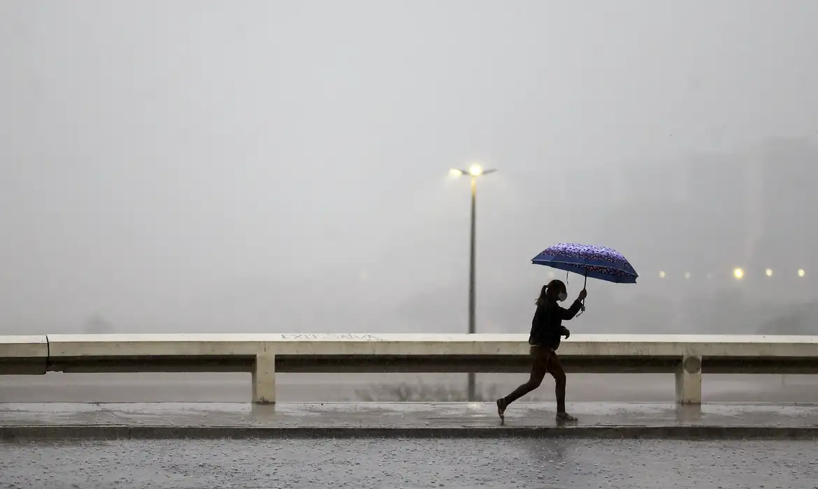 Inmet alerta para fim de semana com chuvas intensas no Norte e Nordeste 2 Plano aberto de uma pessoa caminhando apressada por uma calçada molhada durante um temporal. Ela segura um guarda-chuva azul estampado para se proteger da chuva forte, que cria uma névoa cinzenta ao fundo, escondendo parcialmente a paisagem urbana e postes de iluminação acesos.