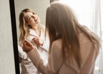 Mulher sorrindo enquanto se olha no espelho, tocando seu cabelo. Tema sobre o hábito de conversar consigo mesma.