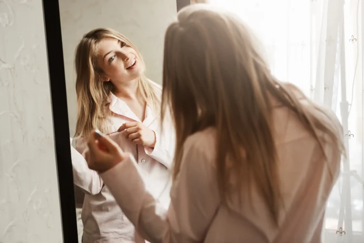 Mulher sorrindo enquanto se olha no espelho, tocando seu cabelo. Tema sobre o hábito de conversar consigo mesma.