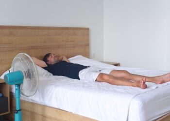 Homem deitado em uma cama de casal branca, com os braços atrás da cabeça, posicionado à frente de um ventilador de mesa azul ligado, em um quarto com cabeceira de madeira.