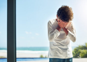 Homem com as mãos cobrindo o rosto, visivelmente triste e angustiada, olhando pela janela, simbolizando o vazio emocional após o Carnaval.