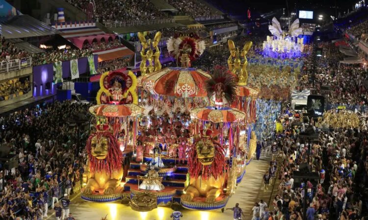Confira a programação das escolas de samba do Carnaval do Rio 2026 1 Vista panorâmica noturna de um desfile no Sambódromo da Marquês de Sapucaí. Em destaque, um grande carro alegórico dourado com esculturas de leões frontais, cercado por componentes da escola de samba em fantasias coloridas sob luzes intensas.