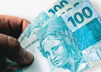 Close de uma mão segurando três notas de 100 reais (cédulas de cor azul com a efígie da República). O fundo é branco e levemente sombreado, focando na textura e nos detalhes das notas de Real.