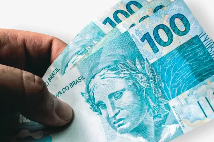 Descubra como consultar se você tem 'dinheiro esquecido' em contas antigas de forma segura 1 Close de uma mão segurando três notas de 100 reais (cédulas de cor azul com a efígie da República). O fundo é branco e levemente sombreado, focando na textura e nos detalhes das notas de Real.