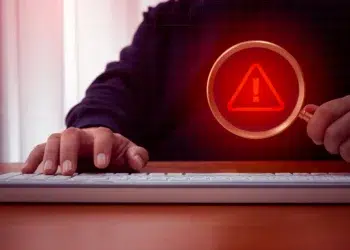 Mão digitando em teclado com lupa exibindo símbolo de alerta vermelho