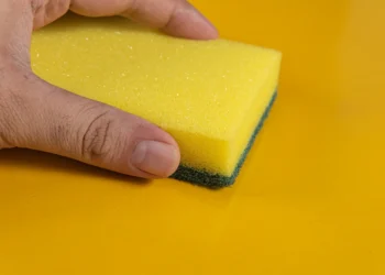 O que acontece se você colocar uma esponja dentro da máquina de lavar? O truque que as lavanderias não contam 4 Esponja amarela sendo segurada, com o foco na parte da esponja e fundo amarelo, ilustra o truque das lavanderias sobre o uso de esponjas na máquina de lavar.