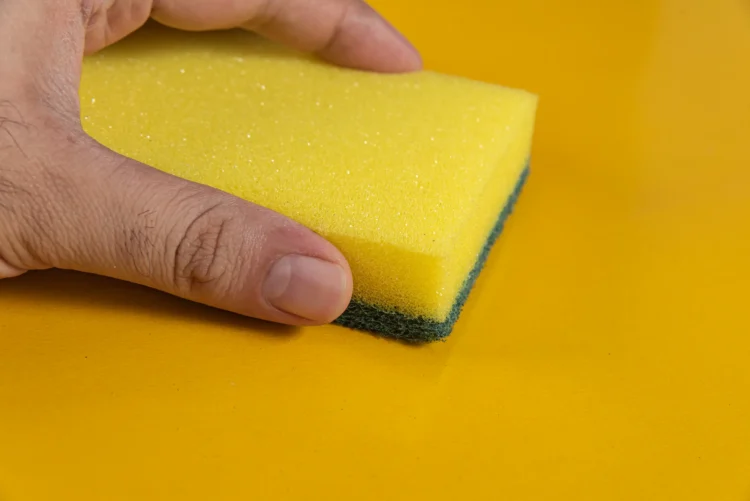 O que acontece se você colocar uma esponja dentro da máquina de lavar? O truque que as lavanderias não contam 1 Esponja amarela sendo segurada, com o foco na parte da esponja e fundo amarelo, ilustra o truque das lavanderias sobre o uso de esponjas na máquina de lavar.