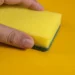 Esponja amarela sendo segurada, com o foco na parte da esponja e fundo amarelo, ilustra o truque das lavanderias sobre o uso de esponjas na máquina de lavar.