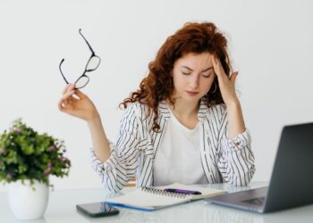 Mulher jovem de cabelos ruivos sentada à mesa de trabalho, segurando óculos e com a mão na testa em sinal de cansaço ou névoa mental diante de um notebook.